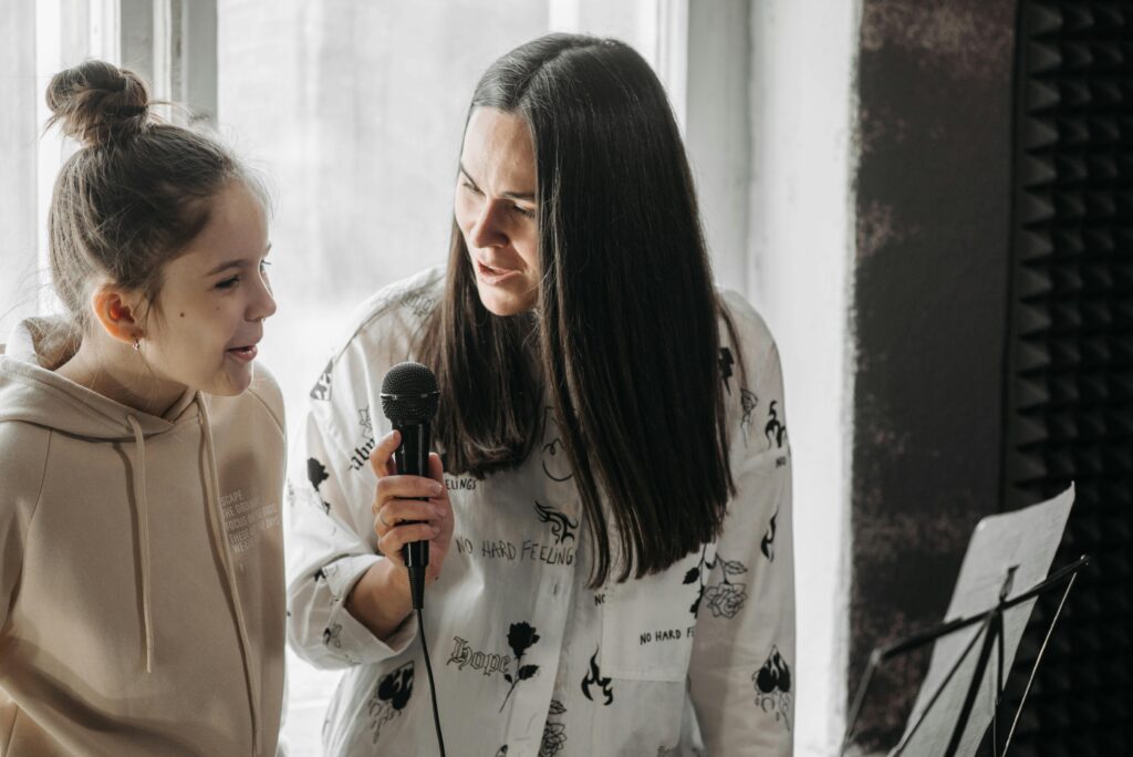 A girl and woman practice singing together indoors using a microphone.